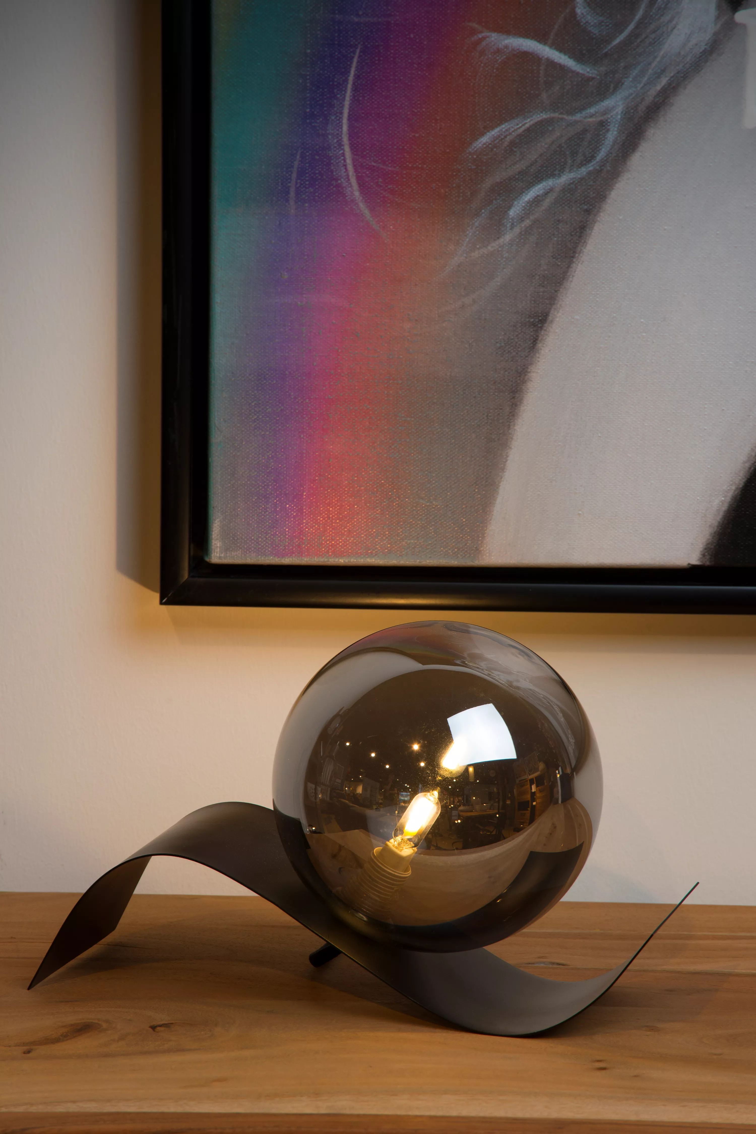 Stolní lampička Yoni se zvlněnou kovovou základnou a koulí z kouřového skla vydává příjemné rozptýlené světlo do obývacího pokoje nebo do ložnice.